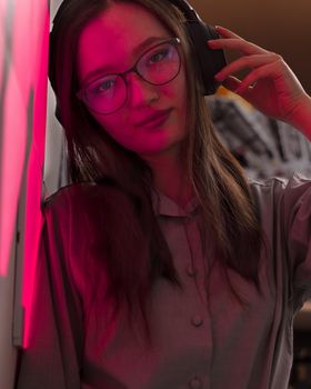 Young smiling woman with wireless headphones in red pink light of neon panels leans on wall looking at lens, nightclub concept, futuristic neon trend of modern electronic music, dark vertical image