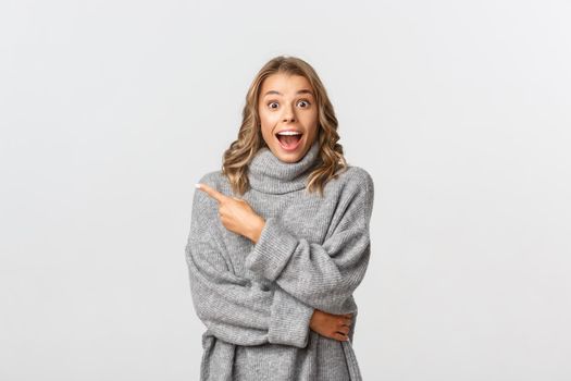 Image of beautiful blond girl in grey sweater pointing finger at upper left corner, showing your logo or banner, looking amazed, standing over white background.