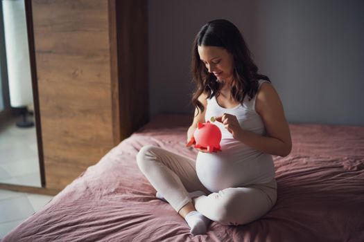 Happy pregnant woman relaxing at home. She is sitting on bed and saving coin in piggy bank.