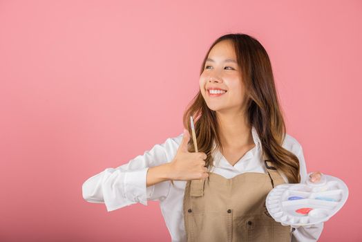 Portrait Asian young woman artist holding brush and paint palette, Happy female painting using paintbrush and palette with colors, studio shot isolated on pink background, Art design workshop