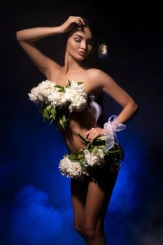 Gorgeous young self assured female model with long dark hair and fresh white flowers covering naked body standing in dark studio - smoke under neon light