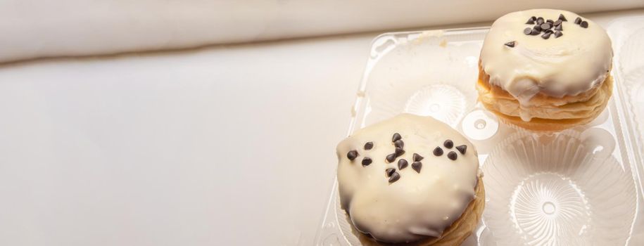 Two delicious donuts with icing sugar and chocolate chips on a white background in a plastic transparent container, a banner with a copy of the space on the left.