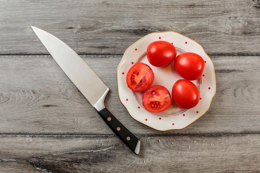 Tomatoes, one of them cut in half on white porcelain plate with small red dots, chef knife lying near. Tabletop view.