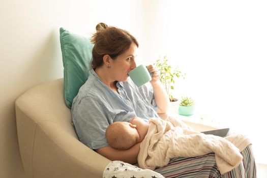 Woman drink tea or coffee during process of breastfeeding of newborn baby. Comfortable breastfeeding