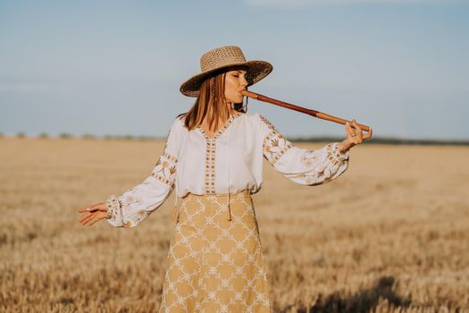 Rural woman plays on wooden flute - ukrainian telenka, tylynka in wheat field. Folk music, sopilka concept. Musical instrument. Musician in traditional embroidered shirt - Vyshyvanka. Quality photo