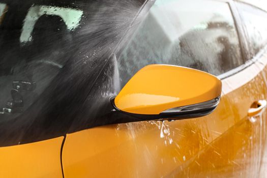 Side mirror of yellow car being washed in self service carwash, water spraying with high pressure, drops flying around.