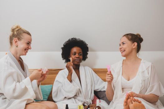 The young women in bathrobes are sitting on the bed and using skincare products