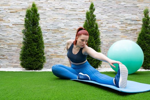young woman with red hair, practising leg stretching exercises on a mat in the garden of her house. Sporty girl training. concept of health and well-being. natural outdoor light.