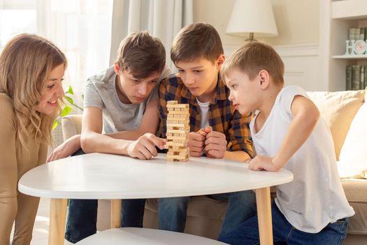 A smiling woman and three boys are enthusiastically playing a board game made of wooden rectangular blocks stacked in the form of a tower in a sunny room. Close-up