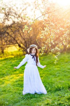 A happy girl in a retro white dress and a hat dancing under flowering trees in the spring, in the garden. Vertical