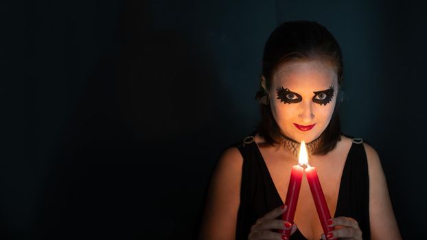 Girl in image of witch looks straight into frame with burning candle in hands on black background. Concept of Halloween and magic. Copy space, banner