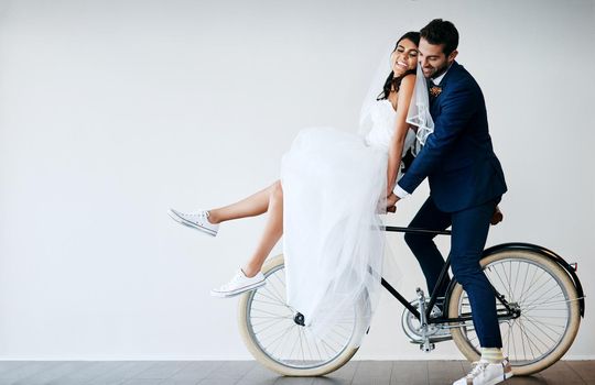 Off to our honeymoon. Studio shot of a newly married young couple riding a bicycle together against a gray background