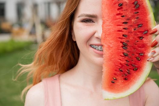 Beautiful red-haired woman smiling with braces on her teeth covers half of her face with a slice of watermelon outdoors in summer.