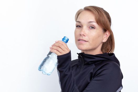 Portrait of caucasian smiling middle aged woman holding plastic bottle of water near mouth to drink on white background looking at camera