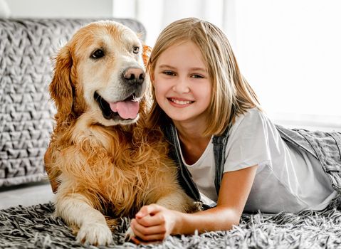 Preteen girl with golden retriever dog lying on floor and smiling. Pretty child kid hugging pet labrador friend indoors