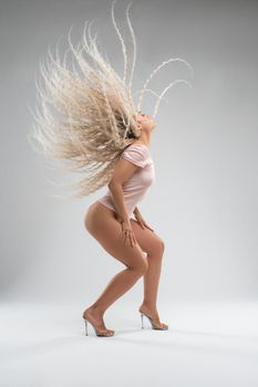 Side view full body of sensual young fit female with long blond curly hair in t shirt and high heeled sandals shaking head while dancing against gray background