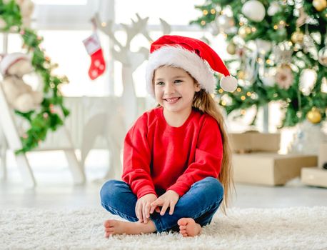 Little girl child wearing Santa hat in Christmas time sitting and smiling at decorated New Year home with festive tree. Cute kid in Xmas time