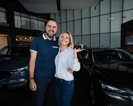 Happy caucasian couple hugging and woman holding new car keys in car showroom