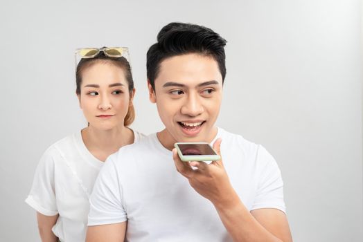 Portrait of a young couple using mobile phones while standing together over gray background, curious woman looking at mans phone
