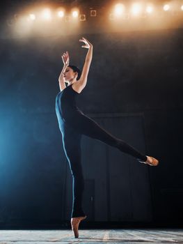 Young beautiful ballerina on smoke stage dancing modern ballet. performs smooth movements with hands against spotlights background. Woman in black costume on scene.