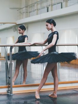 Ballerina in black tutu and pointe stretches on barre in ballet gym. Woman standing near bar and mirror, preparing for perfomance.