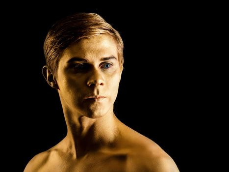 Close up portrait of handsome young ballet dancer with shining golden skin on black background. Body art with gold paint. Fashion style. Man model portrait.