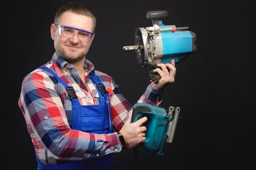 Portrait of smiling young male builder holding tools in both hands while standing and looking at camera on black background.