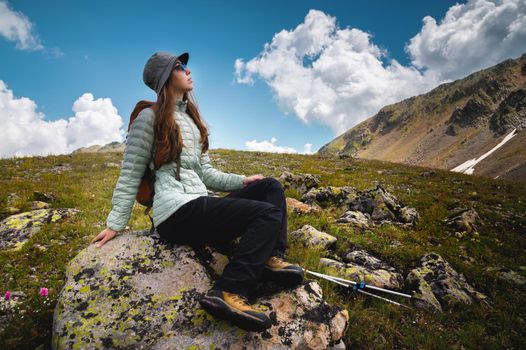 A young woman with a backpack sits on a stone and looks at the sky with clouds and mountains, hiking vacation.