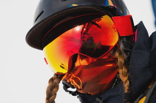 Portrait of a healthy young woman in a sports mask and helmet spending winter holidays in the snowy mountains. Girl rides a lift and looks at the camera.