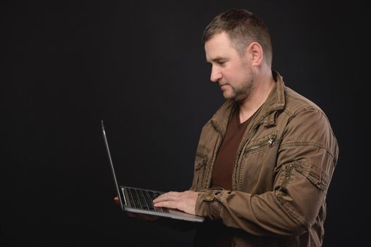 Portrait of an optimistic respectable man in a shirt, standing with a laptop in his hand, looking at the display and working. Studio shot on black background, portrait.