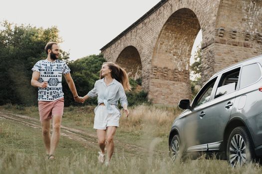 Middle-aged couple running with holding by hands, caucasian man and woman enjoying road trip on SUV car with viaduct on background