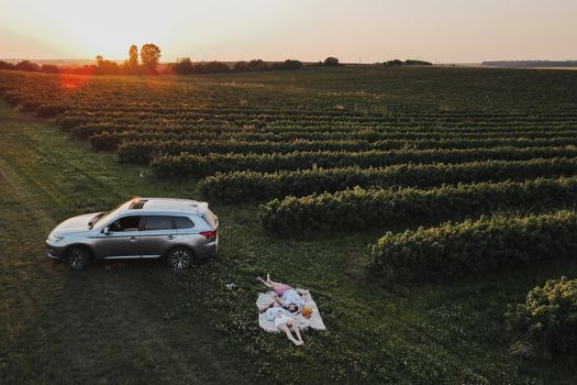 Aerial shot couple enjoying picnic on road trip in the field, man and woman lying on blanket near the SUV car at sunset