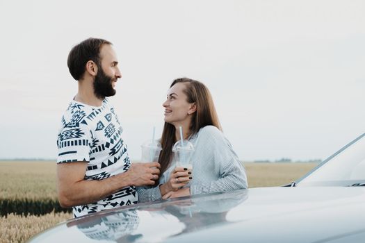 Caucasian cheerful woman and man drinking coffee outdoors near the car in the field, middle-aged couple making stop while on road trip