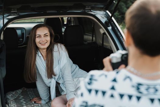 Cheerful caucasian woman sitting in trunk of SUV car while boyfriend photography her on camera