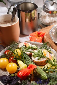 Kitchen garden vegetables on plate with bread, herbs and edible flowers, nasturtium and borage, a cup of coffe with milk on white table, summer breakfast concept, selective focus.