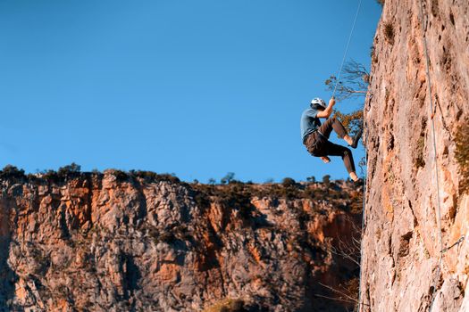 A young man is engaged in rock climbing, mountaineering in a beautiful mountainous area with red rocks, an athlete in a helmet descends from the top against the blue sky on a rope.