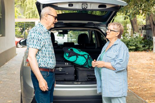 Retired people travelling on vacation by car, talking about urban adventure after loading baggage and luggage in vehicle. Husband and wife preparing to leave on retirement holiday trip.