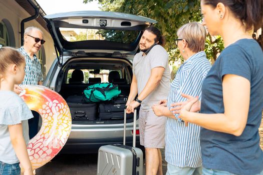 Big family loading baggage in automobile trunk, leaving on holiday vacation journey with parents and grandparents. People travelling on seaside trip with inflatable, luggage and suitcase.