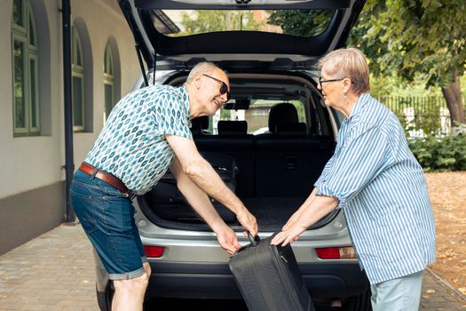 Senior people putting bags in vehicle trunk to leave on retirement holiday trip during summer. Travelling on road trip for recreation, packing baggage and suitcase for urban cityscape vacation.