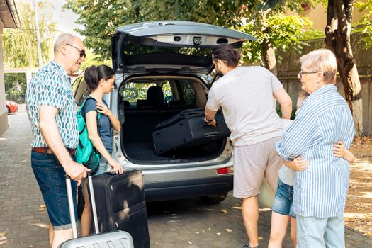 Big european family leaving on vacation journey and loading baggage in automobile trunk. Child travelling with parents and grandparents at seaside during summer, putting luggage in vehicle.