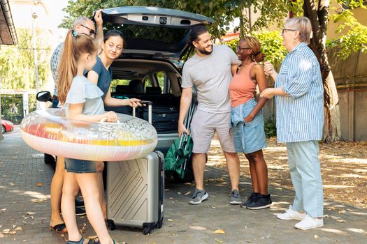 Diverse family and friends travelling on holiday, preparing to leave on summer vacation and loading baggage in car trunk. People sitting in driveway to go to seaside adventure trip.