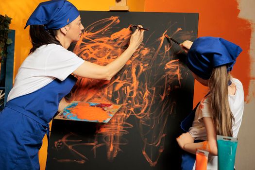 Mother with small kid creating masterpiece with orange color aquarelle paint and dye palette with brush. Little girl learning painting skills on canvas using watercolor and artwork inspiration.