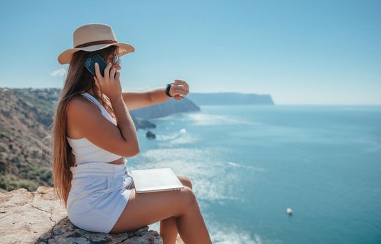 Successful business woman in yellow hat working on laptop by the sea. Pretty lady typing on computer at summer day outdoors. Freelance, travel and holidays concept.