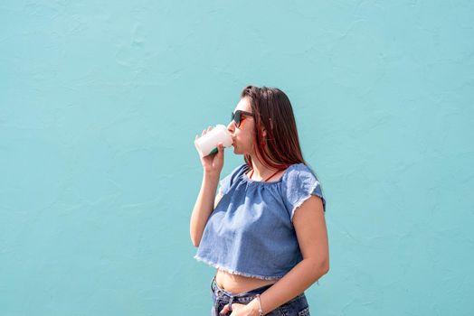 Attractive young woman in summer clothes and sunglasses holding cup of coffee in her hands, dancing on blue wall background at street