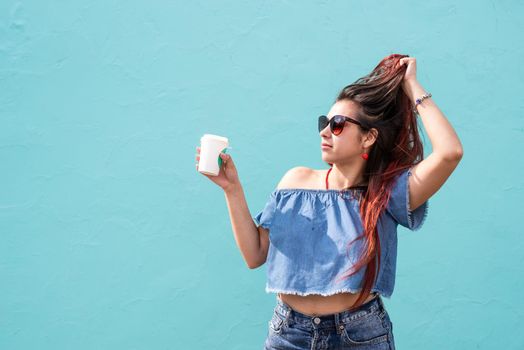 Attractive young woman in summer clothes and sunglasses holding cup of coffee in her hands, dancing on blue wall background at street