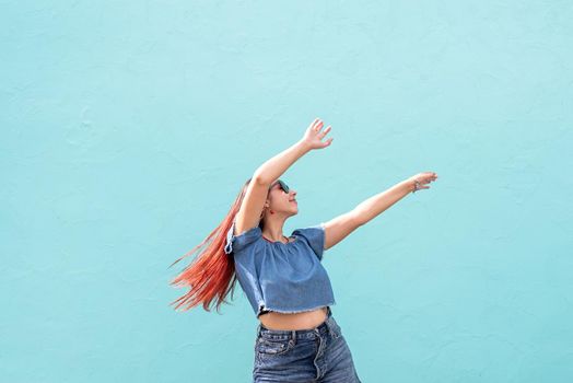 Attractive young woman in summer clothes and sunglasses, dancing on blue wall background at street