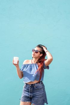 Attractive young woman in summer clothes and sunglasses holding cup of coffee in her hands, dancing on blue wall background at street