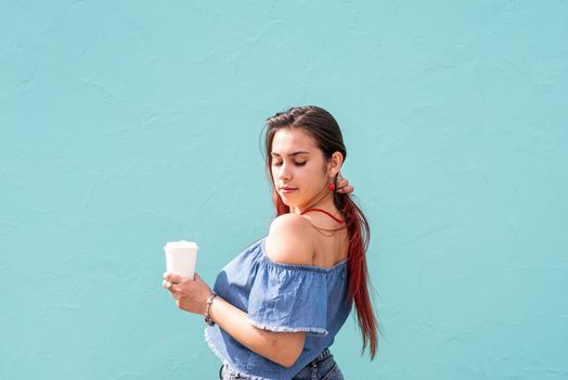 Attractive young woman in summer clothes and sunglasses holding cup of coffee in her hands, dancing on blue wall background at street