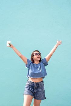 Attractive young woman in summer clothes and sunglasses holding cup of coffee in her hands, dancing on blue wall background at street