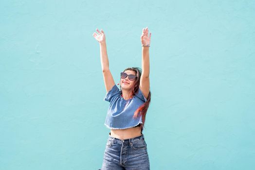 Attractive young woman in summer clothes and sunglasses, dancing on blue wall background at street